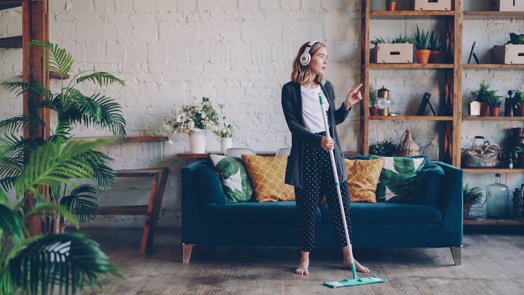 Beautiful young lady is listening to music through wireless headphones and doing housework cleaning the floor with mop. Girl is dancing and singing enjoying cleanup at home.